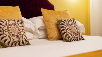 Una cama con sábanas blancas y almohadones decorativos, un cabecero rojo oscuro y una pared amarilla con una lámpara.