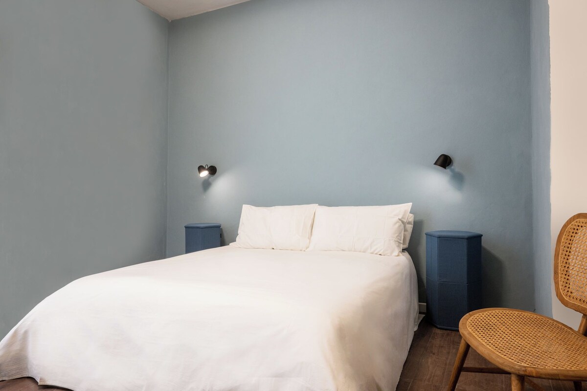 A bedroom with a bed, two wall-mounted lamps, a blue nightstand, and a wicker chair.