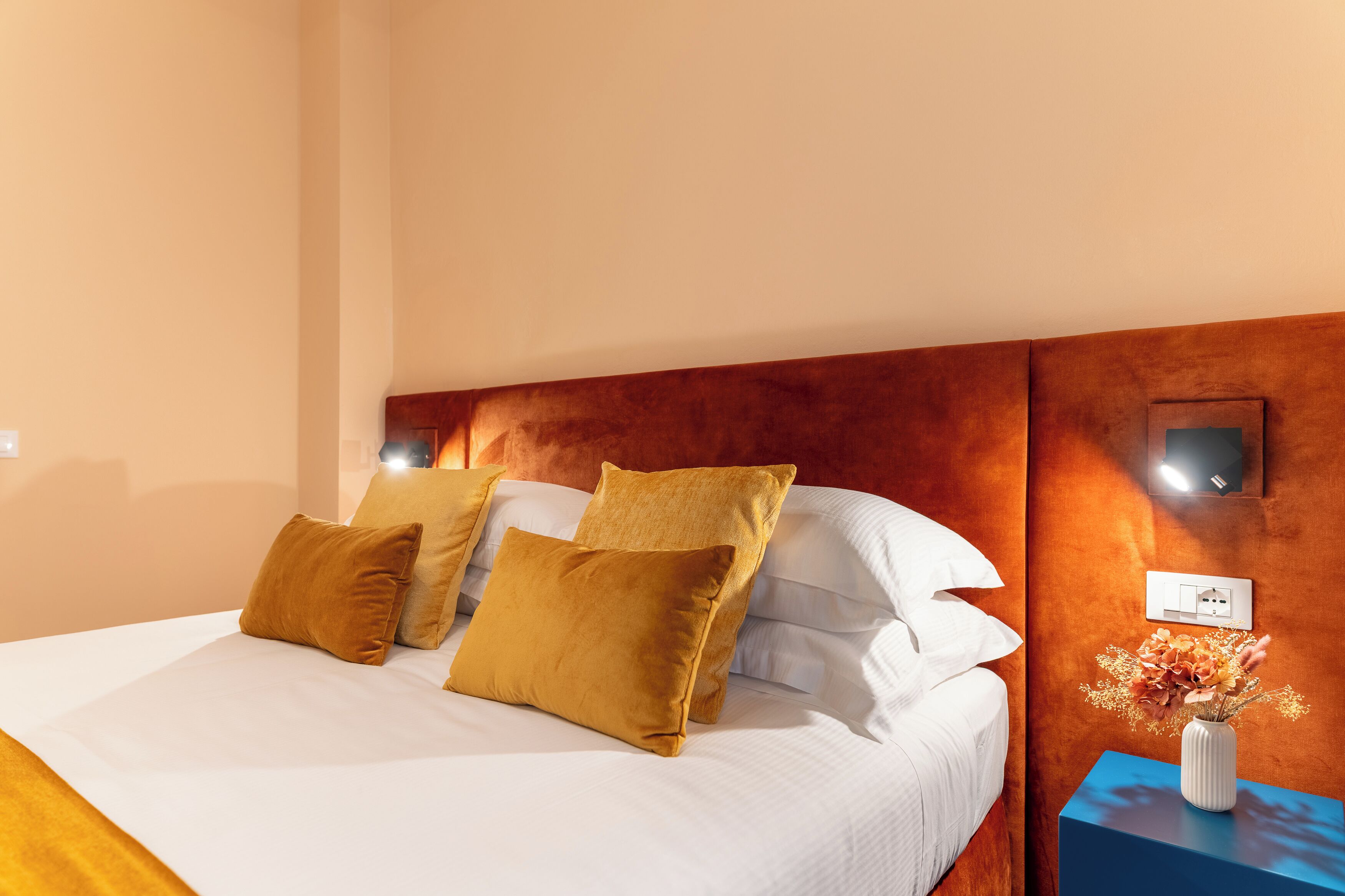 Une chambre d’hôtel avec un lit, des oreillers, une table de chevet, un vase avec des fleurs et une prise électrique au mur.