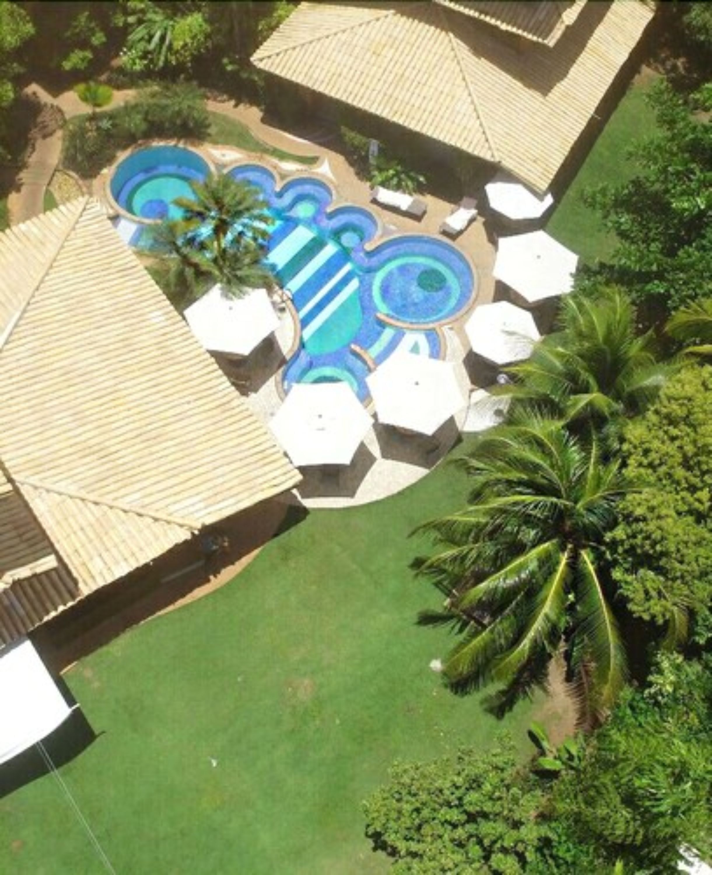 An aerial view of a resort with a central swimming pool, surrounded by lounge chairs and umbrellas, and palm trees.