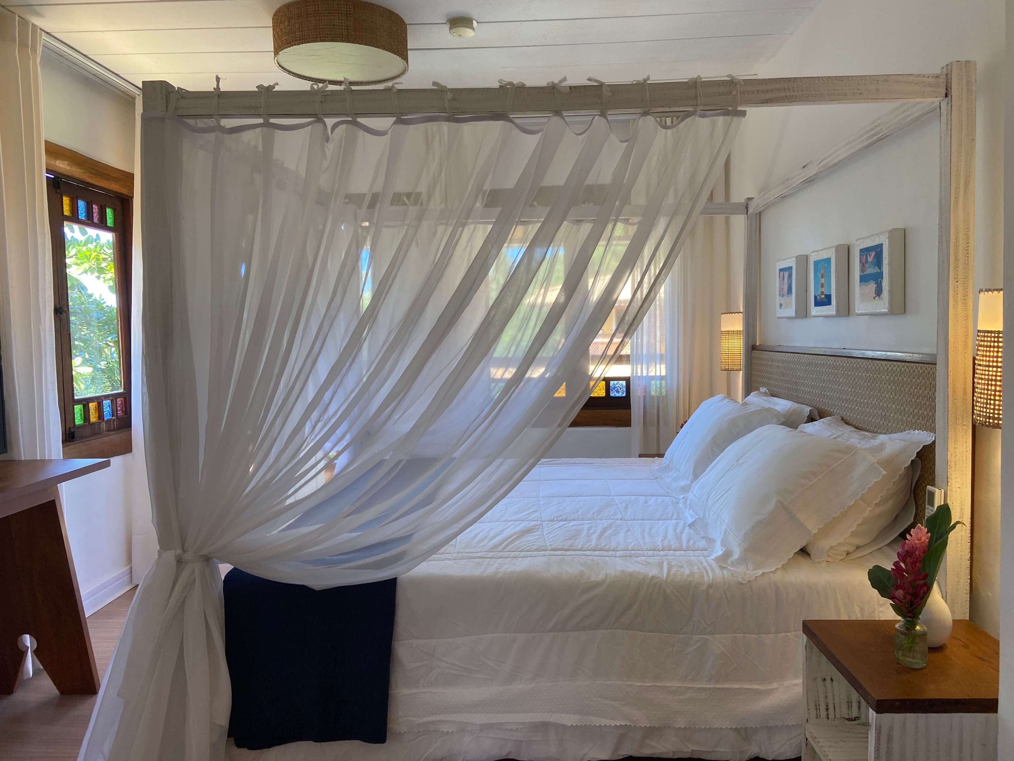 Een slaapkamer met een hemelbed, een nachtkastje met een vaas, en een raam met uitzicht op groen.