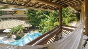 Un balcon avec un hamac, une piscine et une vue sur un bâtiment doté d’un toit en tuiles et d’une végétation luxuriante.
