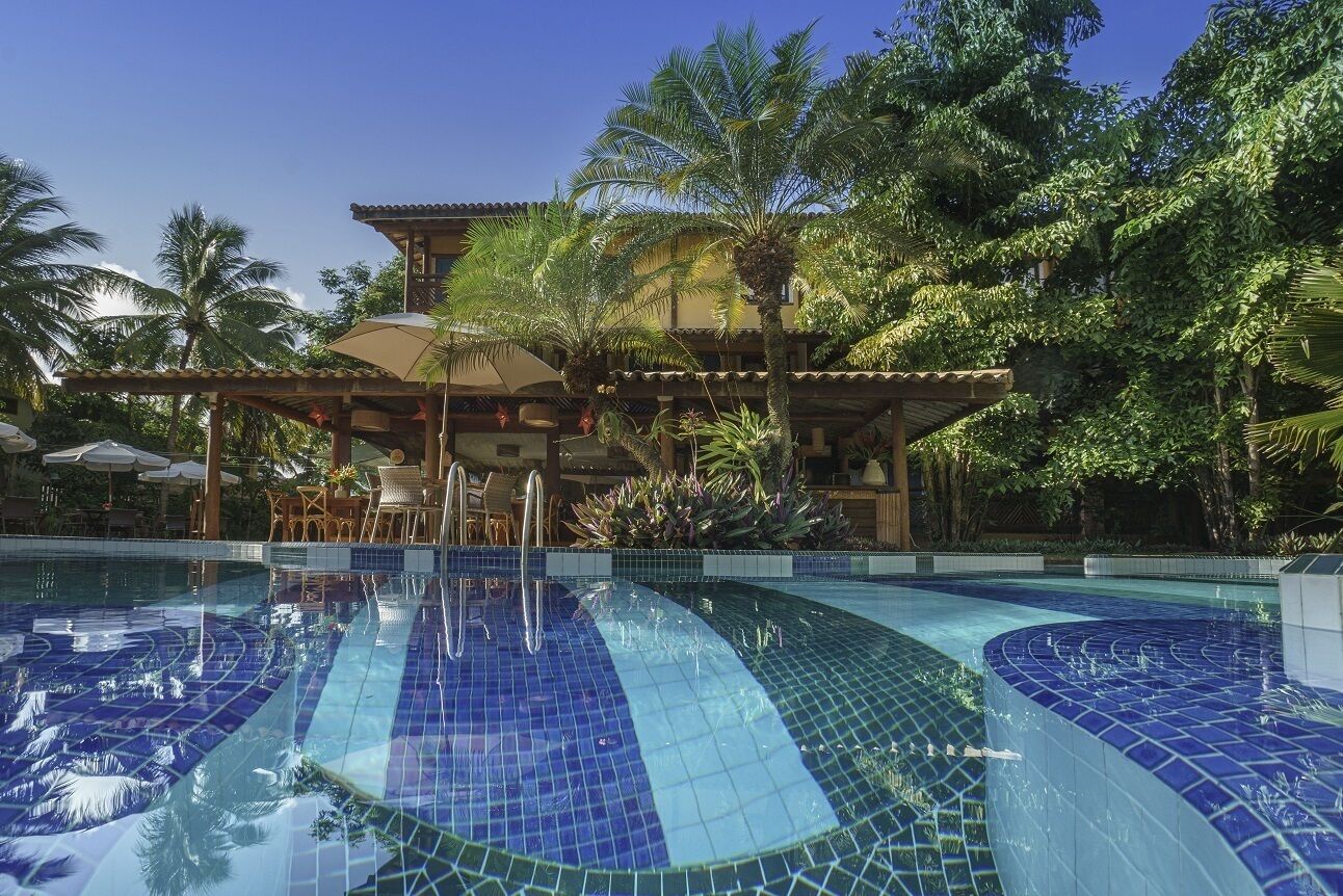 Una piscina con mosaicos azules, rodeada de palmeras y una zona de estar techada.