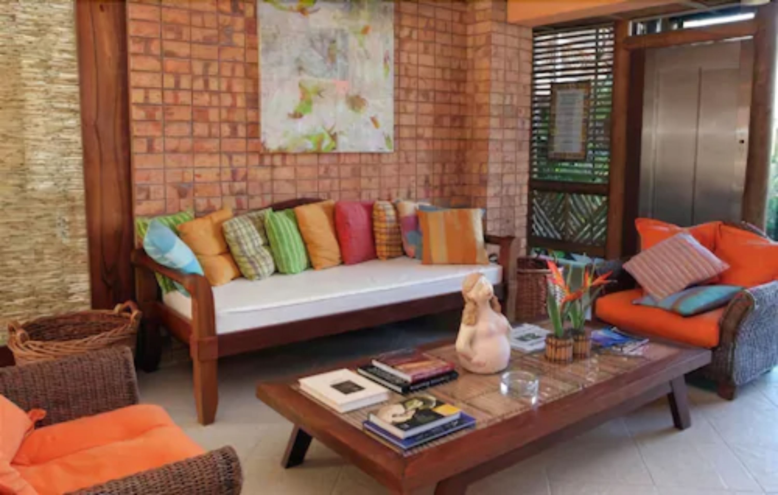 A cozy living area with a sofa and armchairs, a coffee table with books and decorative items, and a brick wall with a colorful painting.