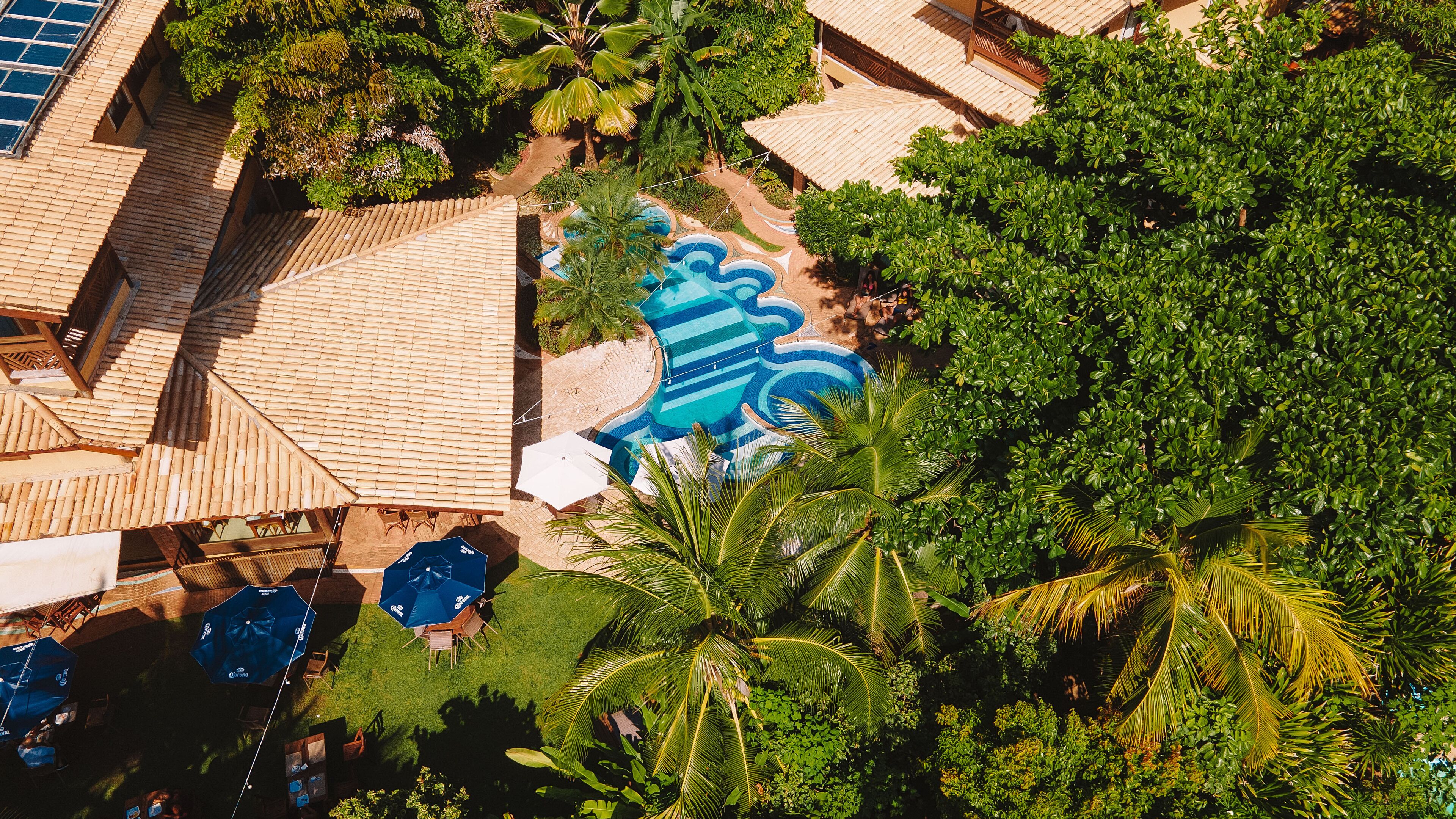 Luchtbeeld van een resort met bamboibedekkingen, een zwembad met een blauwe muurschildering en weelderige begroeiing.
