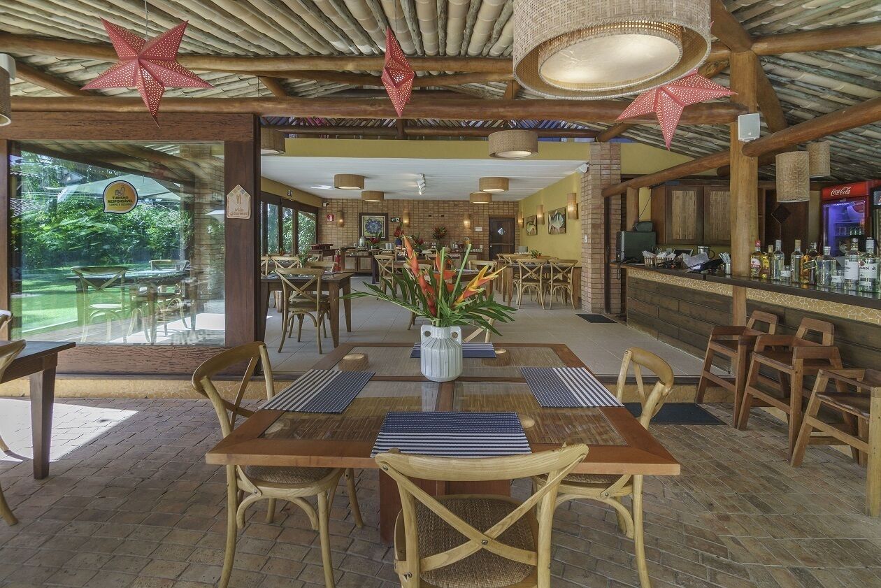A restaurant with wooden tables and chairs, a bar area, and large windows overlooking a garden.
