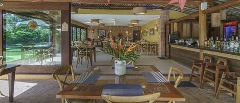 A restaurant with wooden tables and chairs, a bar area, and large windows overlooking a garden.