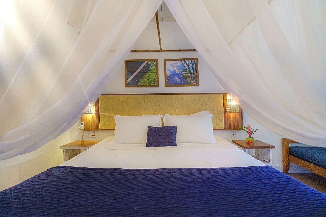 A bedroom with a bed, white curtains, two wall-mounted lamps, and two framed pictures.
