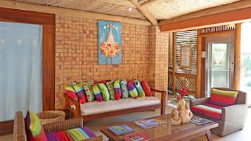 Um quarto aconchegante com parede de tijolos, teto de madeira, um banco estofado com almofadas coloridas, cadeiras de vime e uma mesa de centro de madeira com livros.