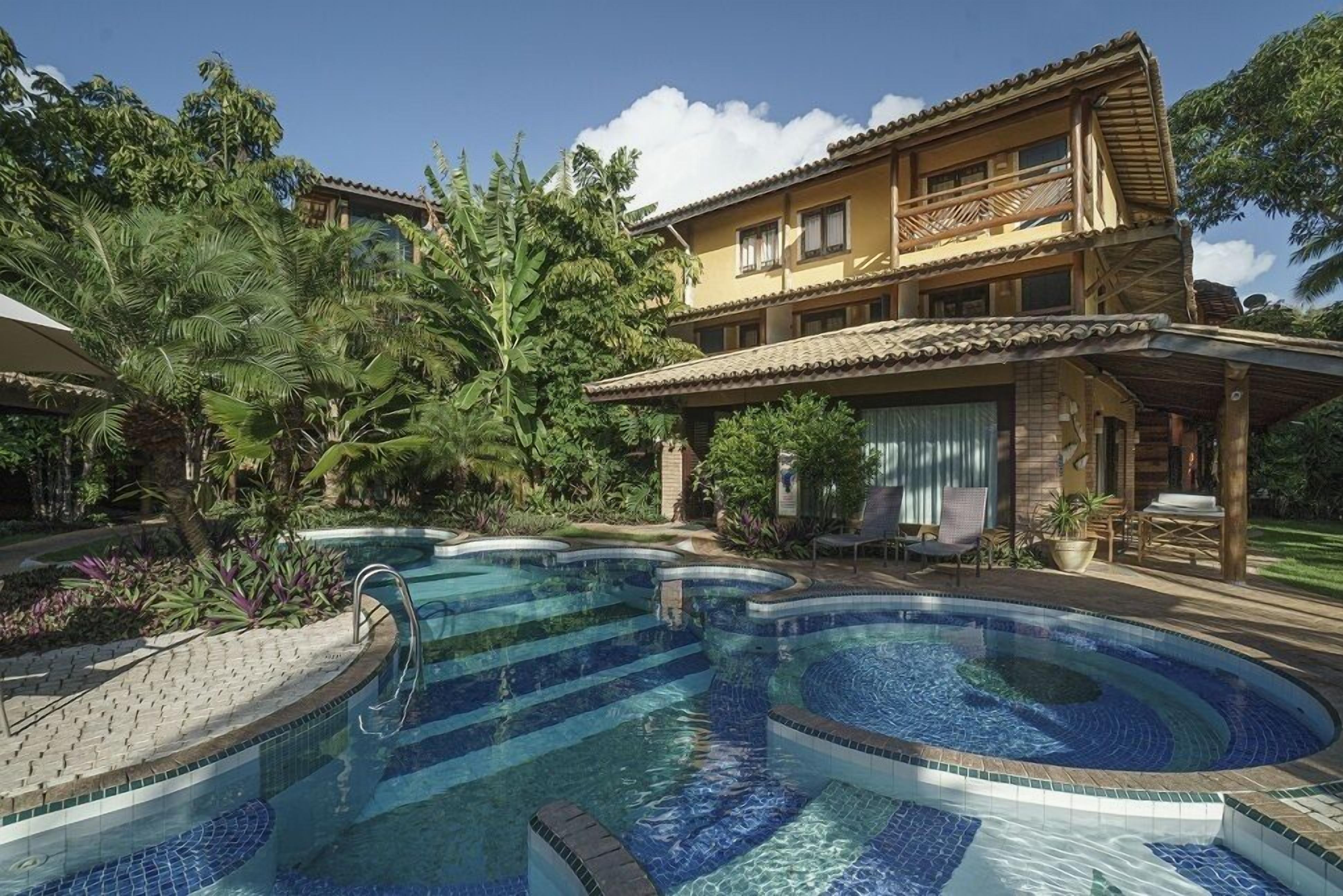 A pool area with a two-story building, surrounded by lush greenery and a clear blue sky.