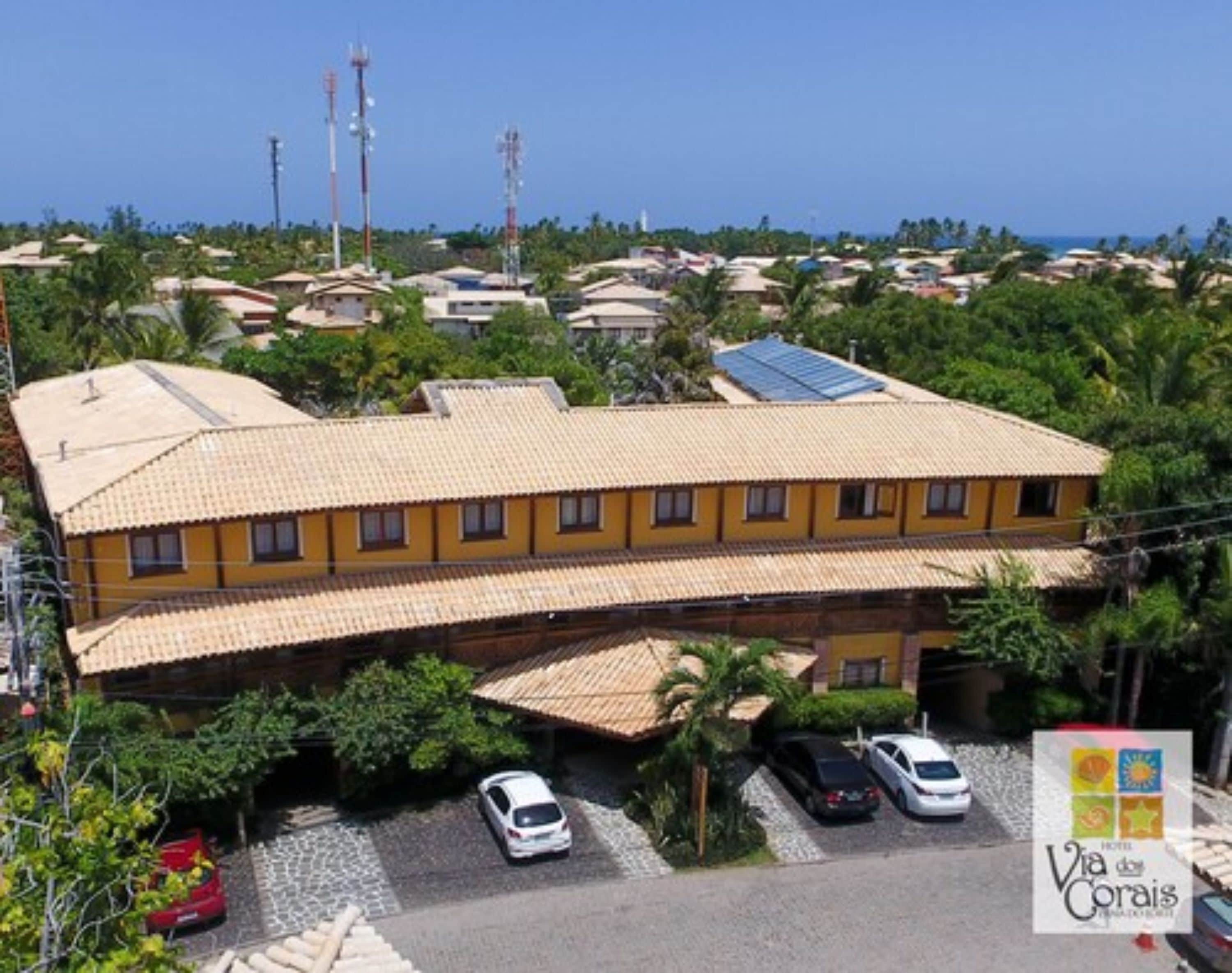 Um hotel com telhado de azulejos, vários carros estacionados na entrada e vista clara do bairro ao redor.