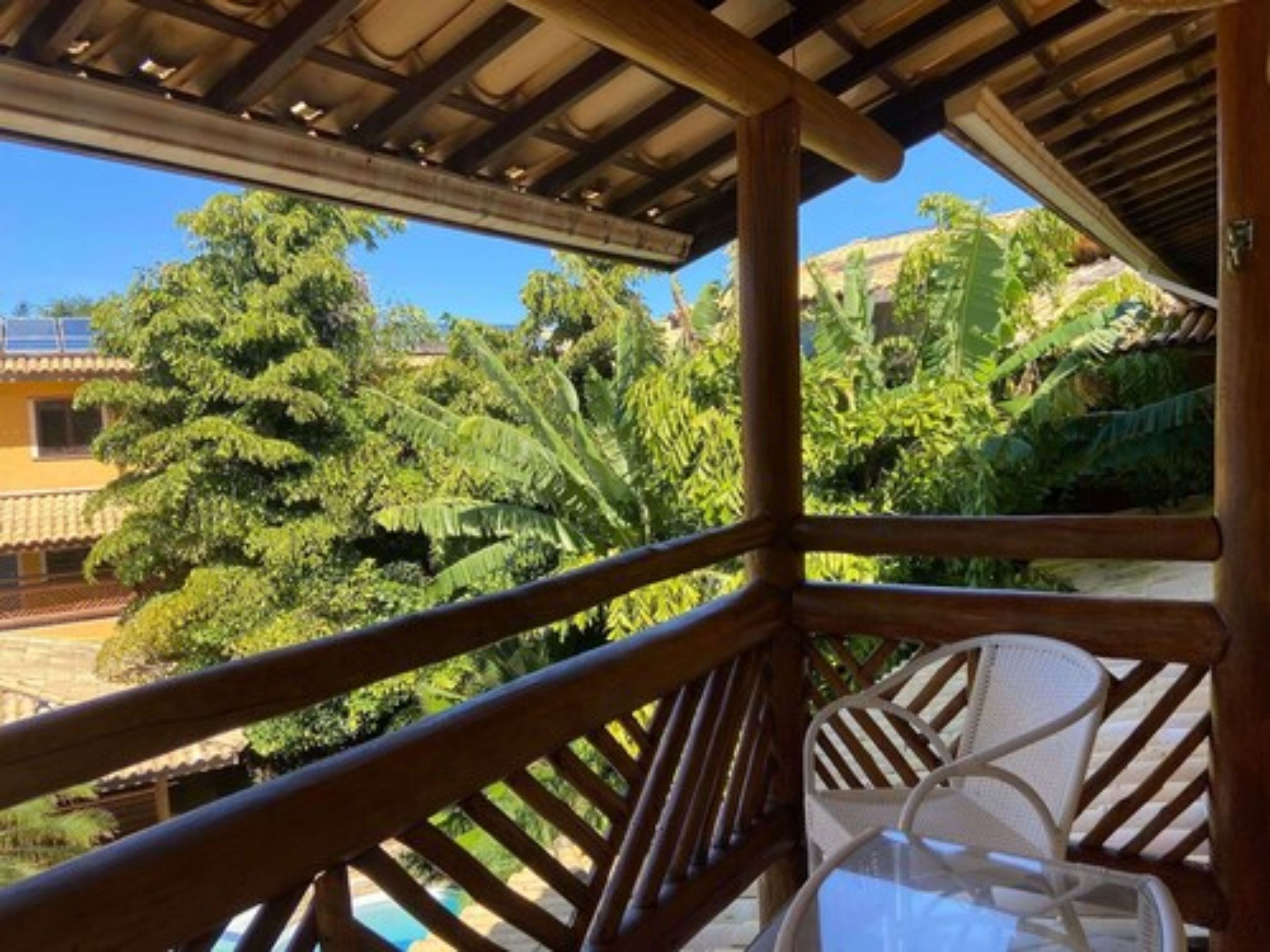 A wooden balcony with white chairs, overlooking a lush garden and a pool.