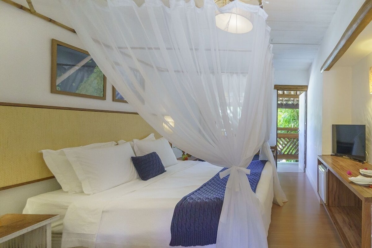 Une chambre à coucher avec un lit sous un dais, un téléviseur posé sur une table en bois et une vue sur la verdure à l’extérieur.