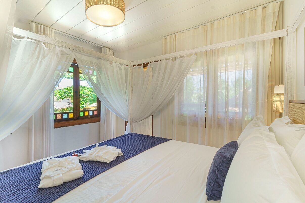 A hotel room with a large bed, white curtains, and a view of greenery outside.