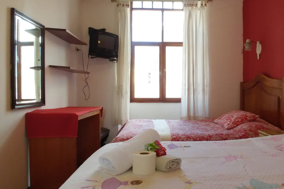 Un dormitorio con cama, un escritorio pequeño, televisor montado en la pared y ventana con cortinas.