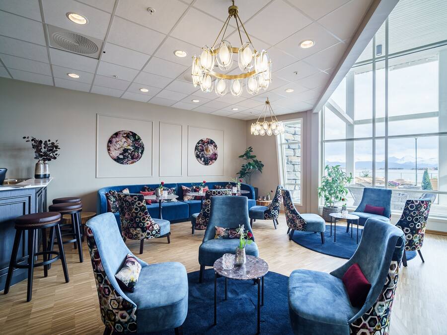 A modern lounge area with a blue sofa set, patterned chairs, a bar area, and large windows offering a view.