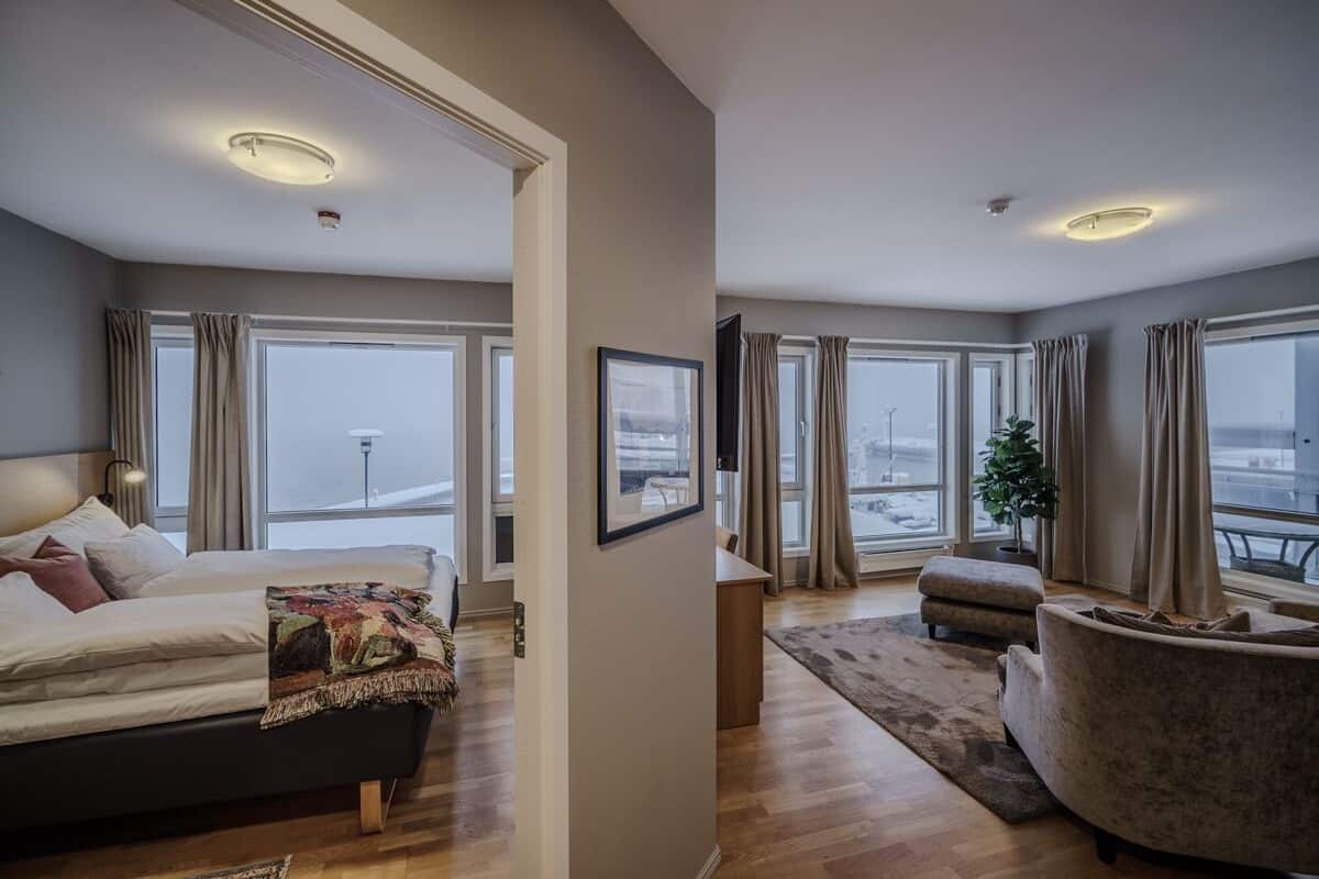 A modern hotel room with a large bed, a sofa, a small table, and a view of the city through large windows.