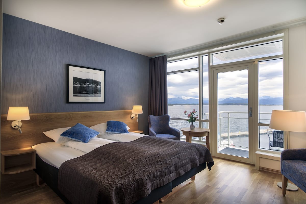 A hotel room with a large bed, two bedside tables with lamps, a chair, a small table with a vase, and a large window with a view of the water and mountains.