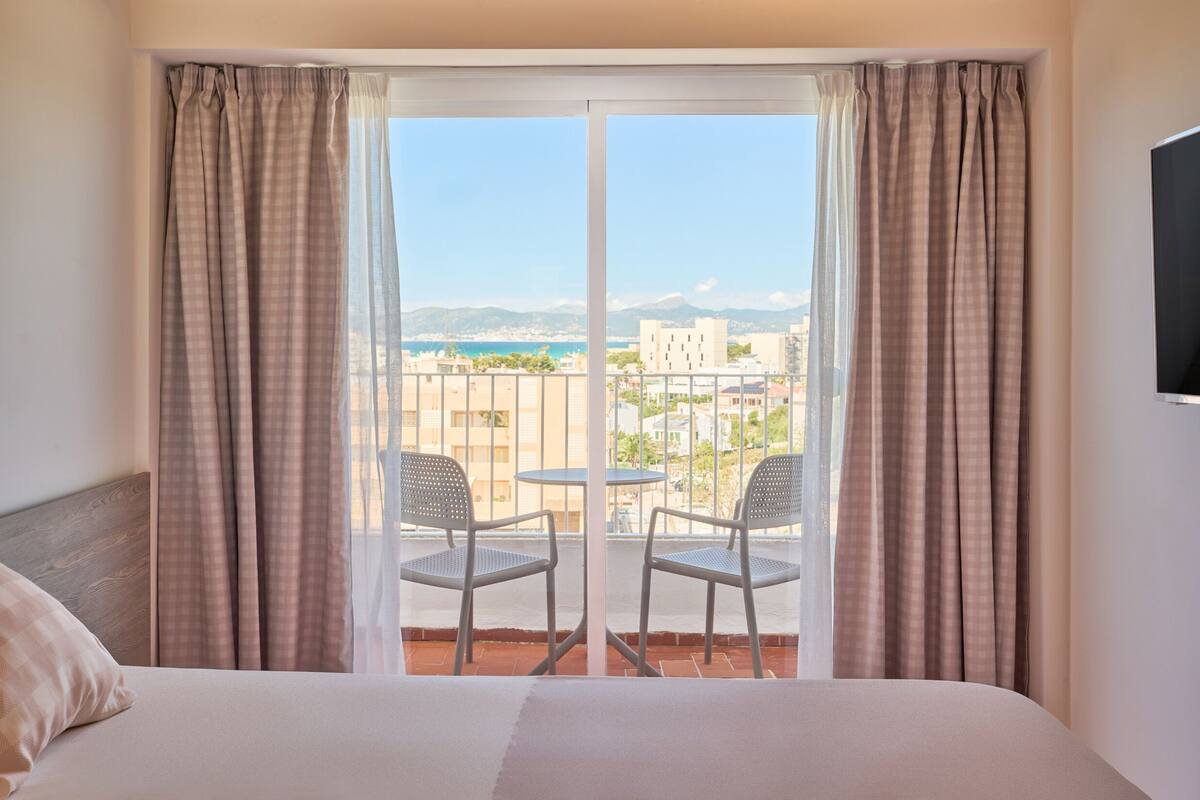 A hotel room with a bed, a balcony with two chairs, and a view of the city and sea.