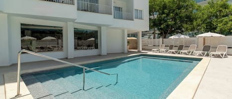 A modern hotel with a rectangular swimming pool, outdoor seating with umbrellas, and a multi-story building with balconies.