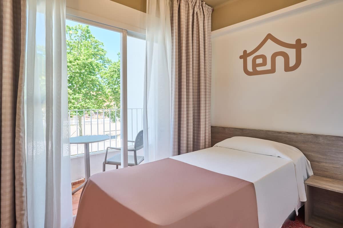 A hotel room with a bed, a small table, and a balcony with a chair.