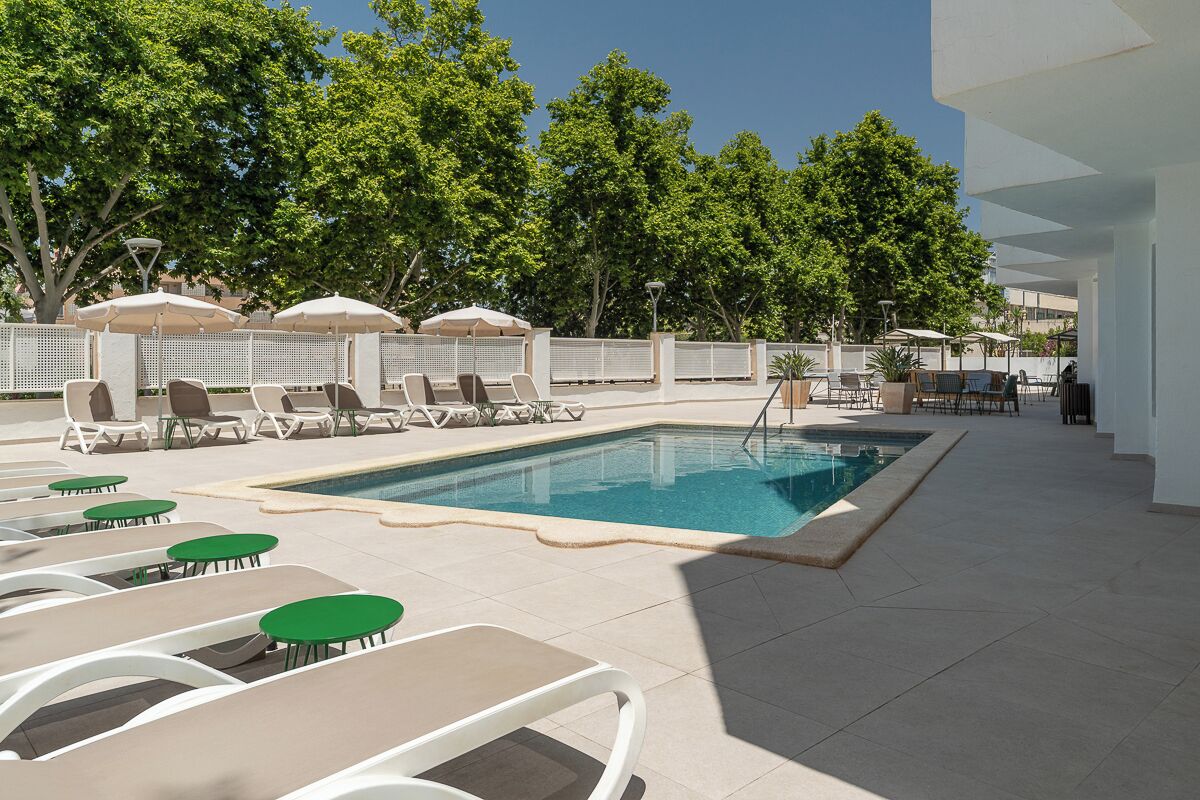 A poolside area with lounge chairs and tables, a swimming pool, and a white building in the background.