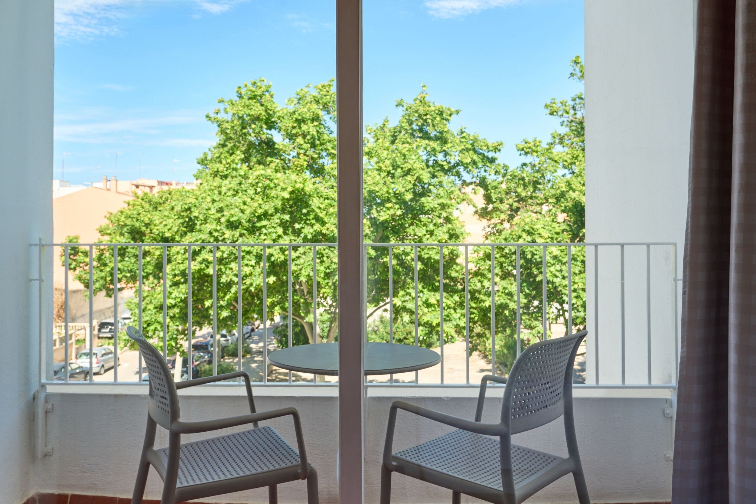 A balcony with two chairs and a table, overlooking a street with parked cars and trees.