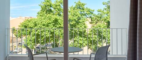 A balcony with two chairs and a table, overlooking a street with parked cars and trees.