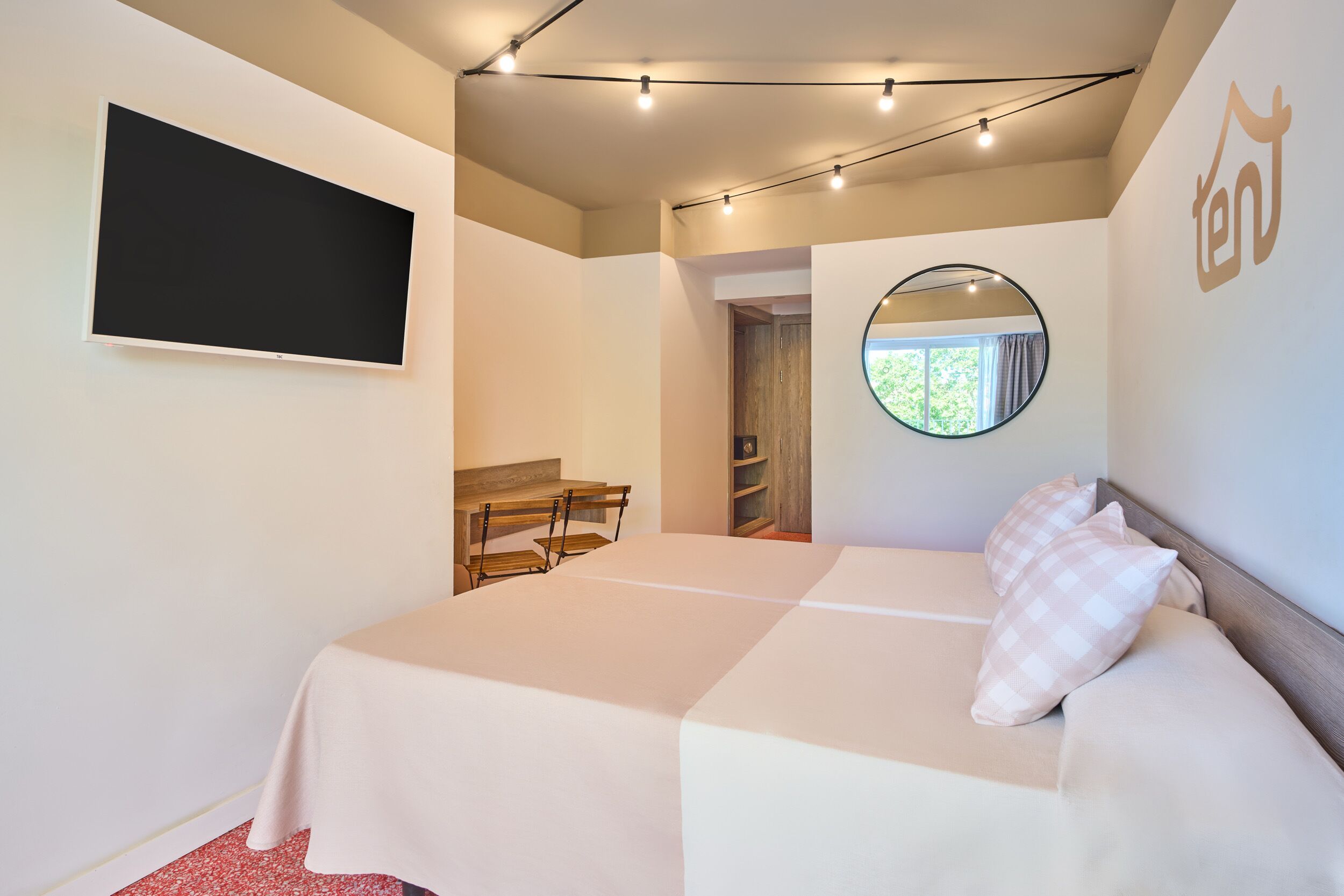 Un dormitorio con una cama, un televisor montado en la pared, un espejo redondo y una pequeña mesa con sillas.