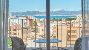 Un balcón con mesa y sillas, con vistas a un paisaje urbano costero que incluye edificios y montañas.