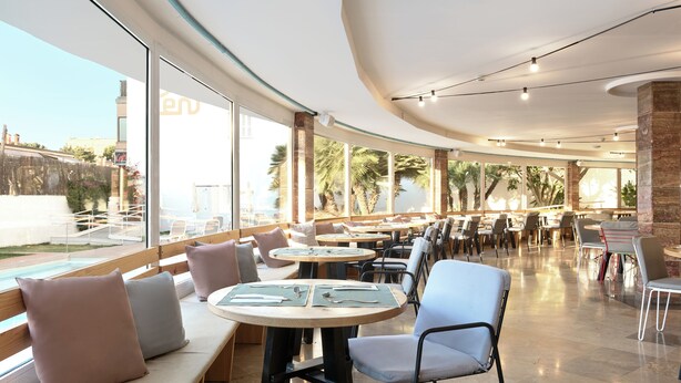 Un comedor amplio con mesas y sillas redondas, grandes ventanales que ofrecen vistas a palmeras y una piscina.