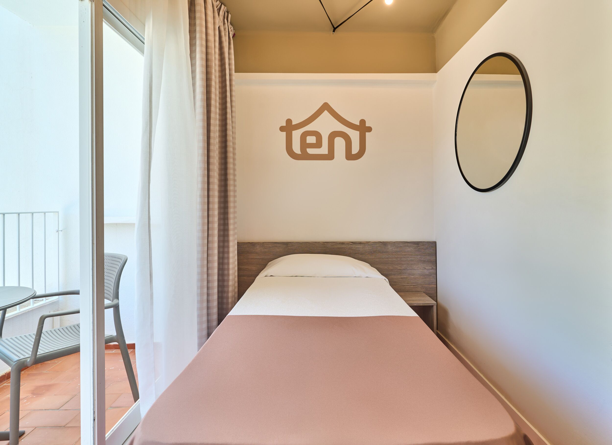 Ein Einzelbett mit Kopfteil, ein an der Wand befestigter Spiegel und ein Fenster mit Vorhängen.