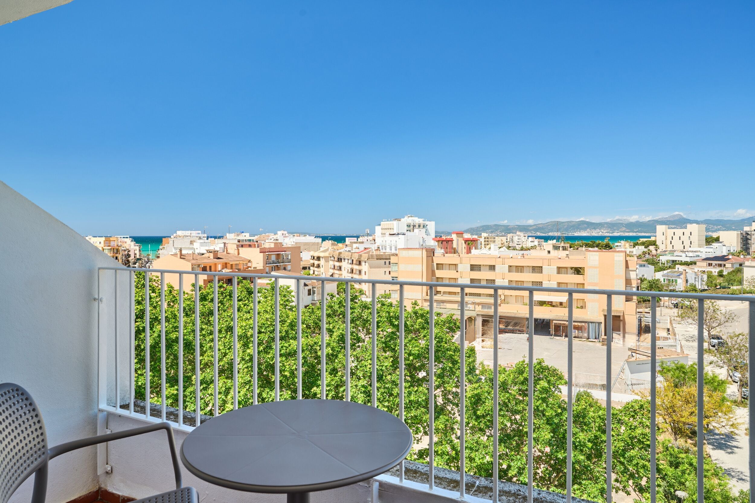 Un balcone con tavolo e sedie, affaccia su un panorama urbano con edifici e cielo sereno.