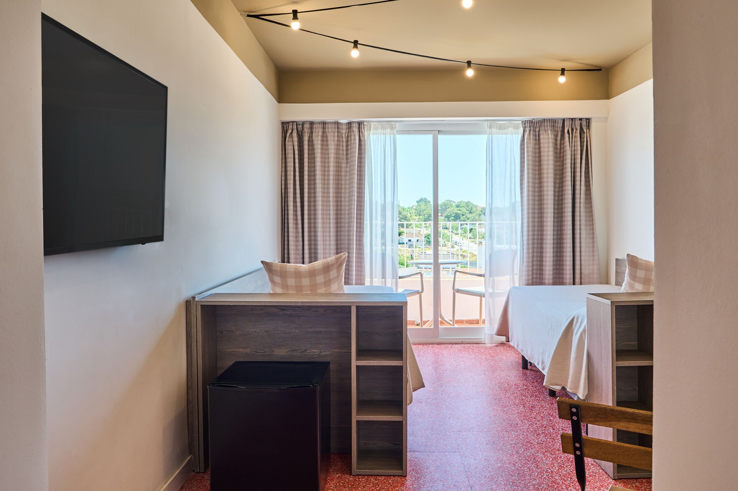 A bedroom with a bed, a desk, a chair, a TV, and a balcony with curtains.