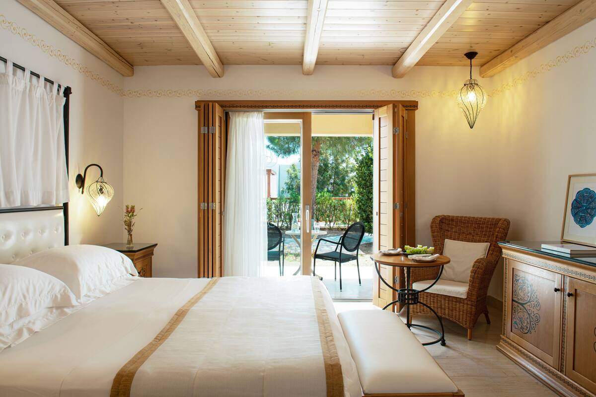 Ein Hotelzimmer mit einem großen Bett, einem Esstisch mit Stühlen, einem Korbstuhl, einem Schrank und Blick ins Freie.