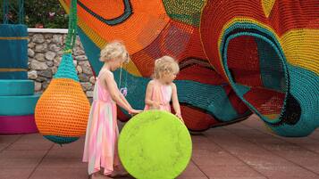 Two children holding a large green object and a hanging orange and green object in an outdoor setting with a colorful, abstract wall.