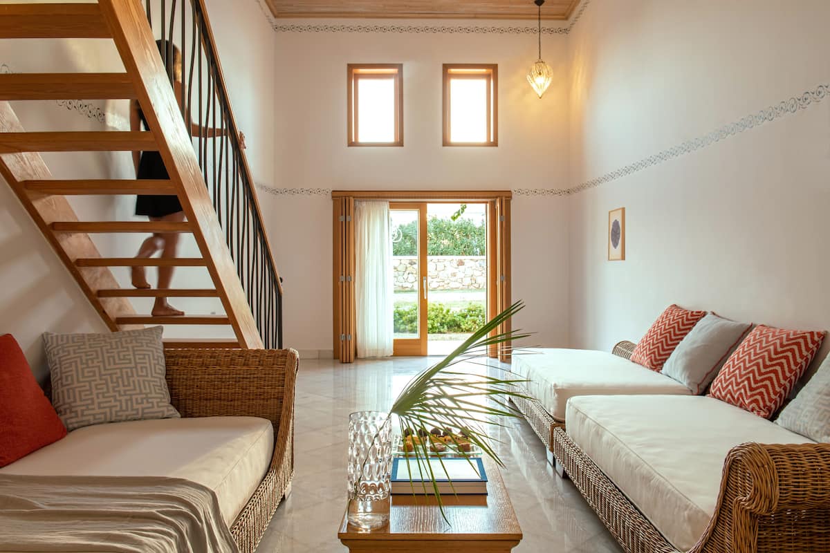 Ein geräumiger Wohnbereich mit einer Holztreppe, zwei Sofas mit gemusterten Kissen, einem Couchtisch mit einem Buch und einer Pflanze sowie einem Blick durch eine Glastür in einen Außenbereich.