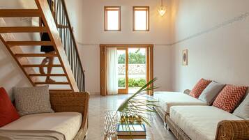 Amplia sala de estar con una escalera de madera, dos sofás con cojines estampados, una mesa de centro con un libro y una planta, y vistas a un espacio exterior a través de un ventanal.