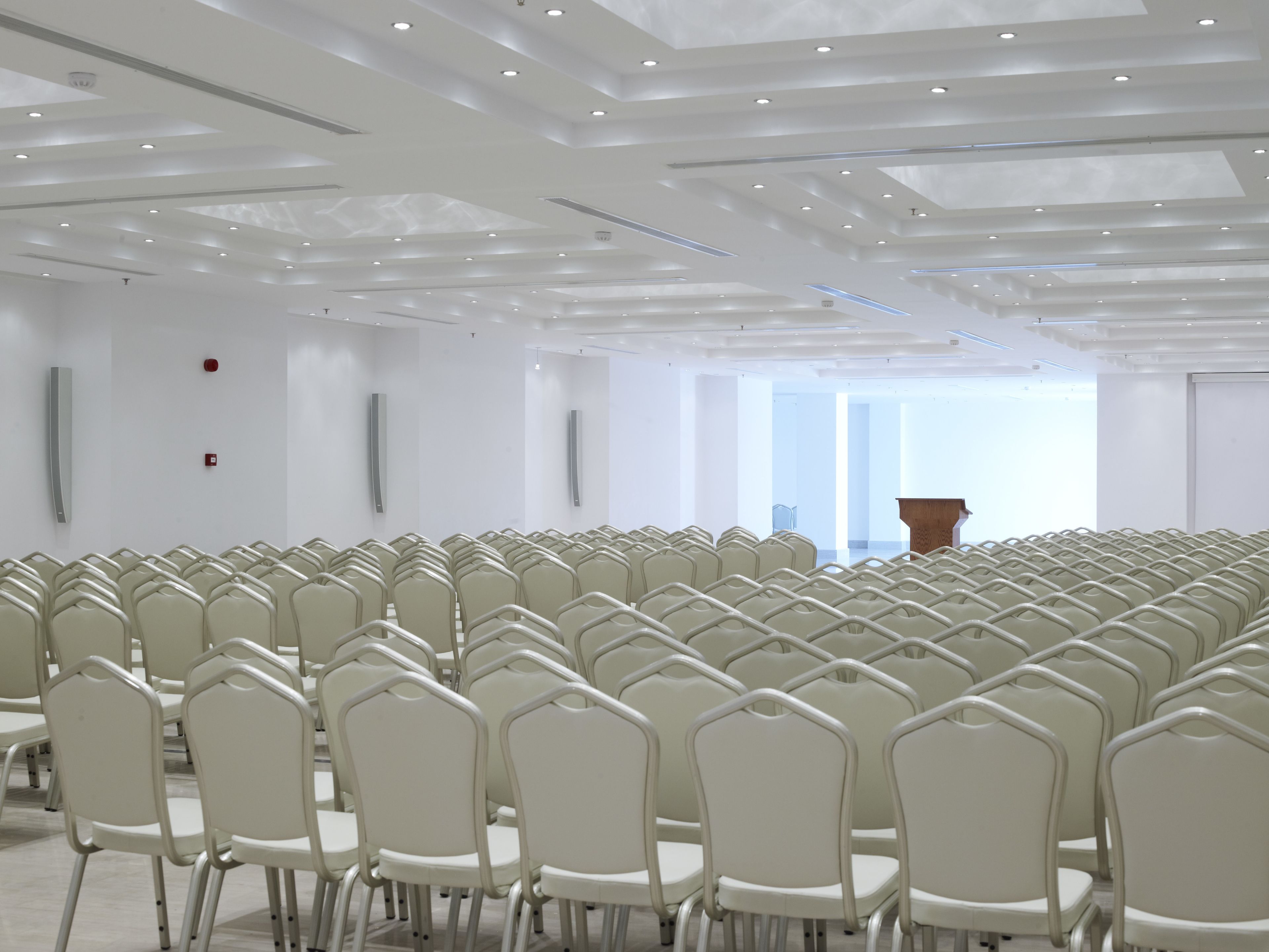 Un salón de conferencias con filas de sillas blancas, un atril y un techo alto con iluminación empotrada.