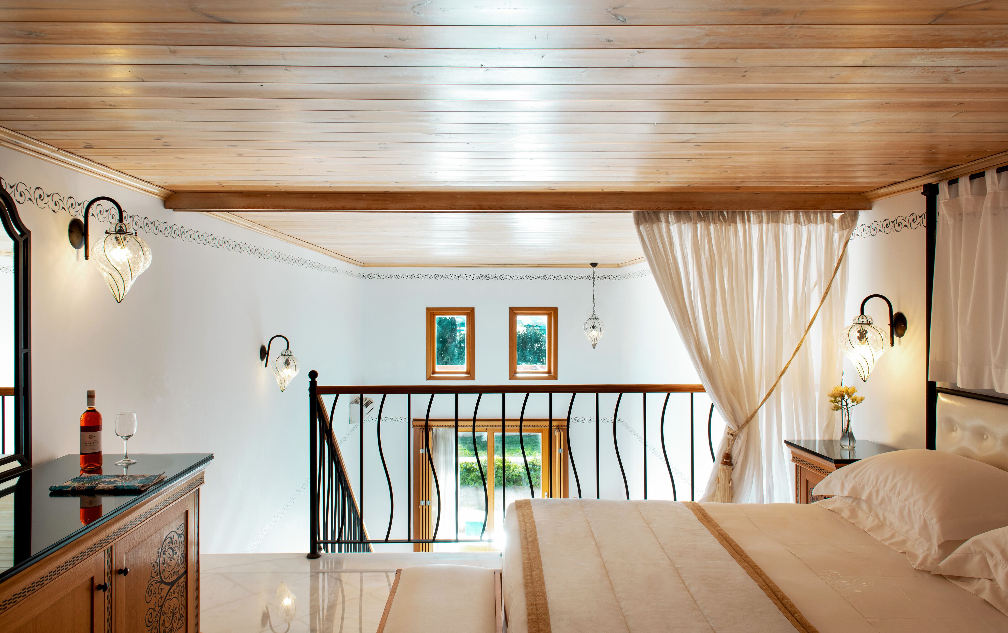 Quarto com teto de madeira, cama com roupa de cama branca, mesa de cabeceira com abajur e vista para o verde lá fora.