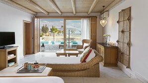 Amplia habitación con una cama grande, un sofá de mimbre, televisión y vistas a una piscina y palmeras.