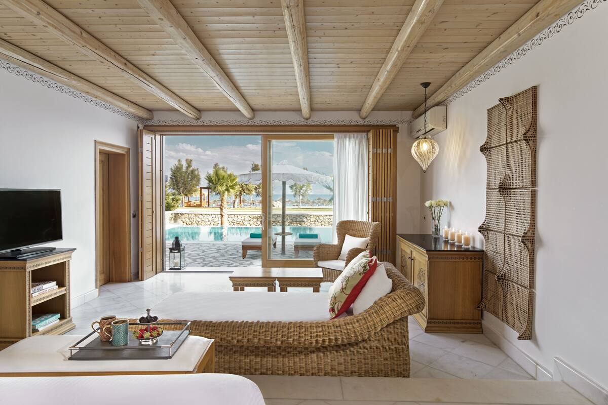 Ein geräumiges Zimmer mit einem großen Bett, einem Korbsessel, einem Fernseher und Blick auf einen Pool und Palmen.