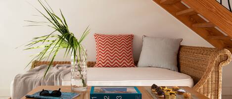 Una habitación acogedora con una mesa de madera, un sofá de mimbre, una cama blanca y un cojín rojo y blanco.
