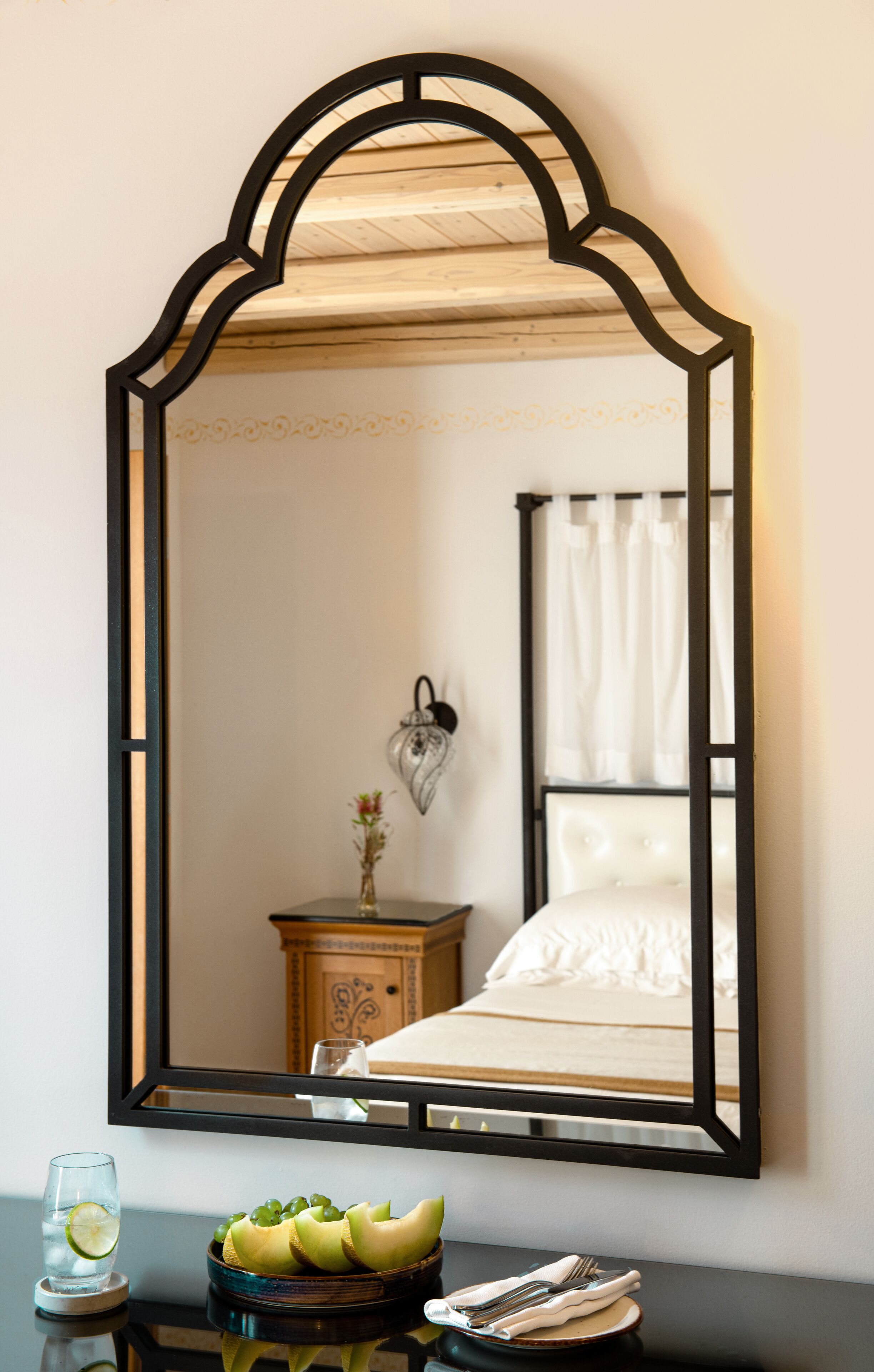 Une chambre à coucher avec un grand miroir, une table de chevet ornée d’un vase et de fruits, et un lit recouvert de linge de lit blanc.
