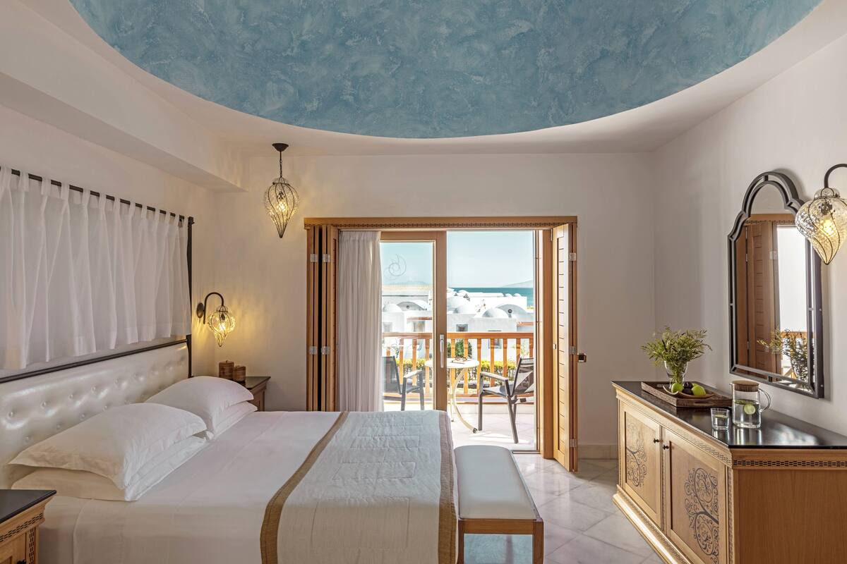 Ein Schlafzimmer mit Bett, Nachttisch, Schminktisch, Kommode, Spiegel und einem Balkon mit Meerblick.