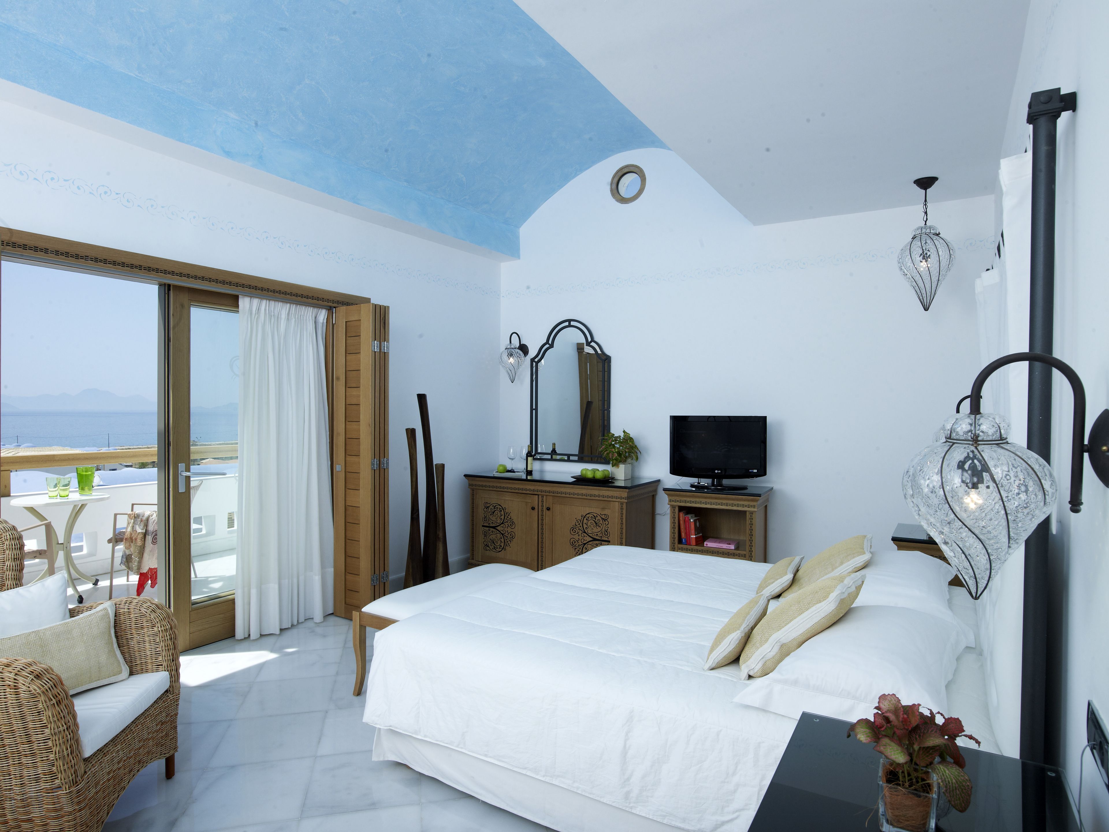 A bedroom with a bed, a TV, a dresser, and a balcony with a view of the sea.