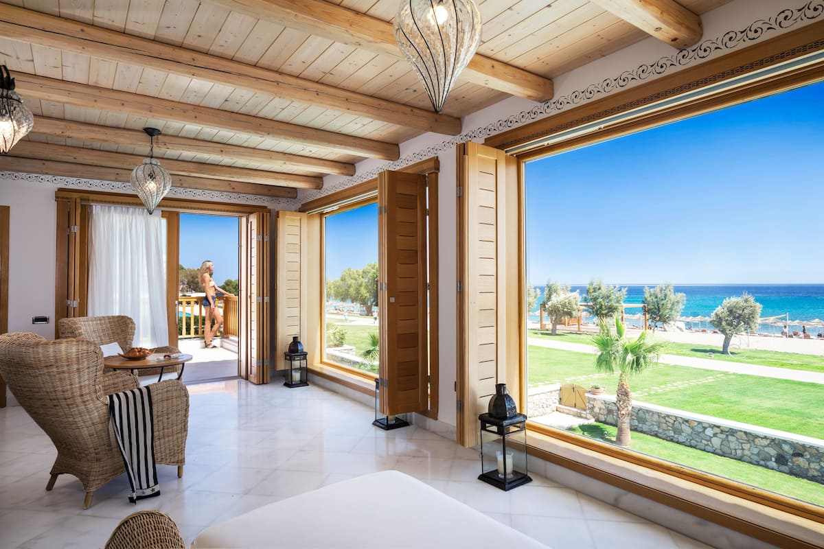 Ein geräumiges Zimmer mit großem Fenster und Meerblick, Korbmöbeln und einer Decke mit Holzbalken.