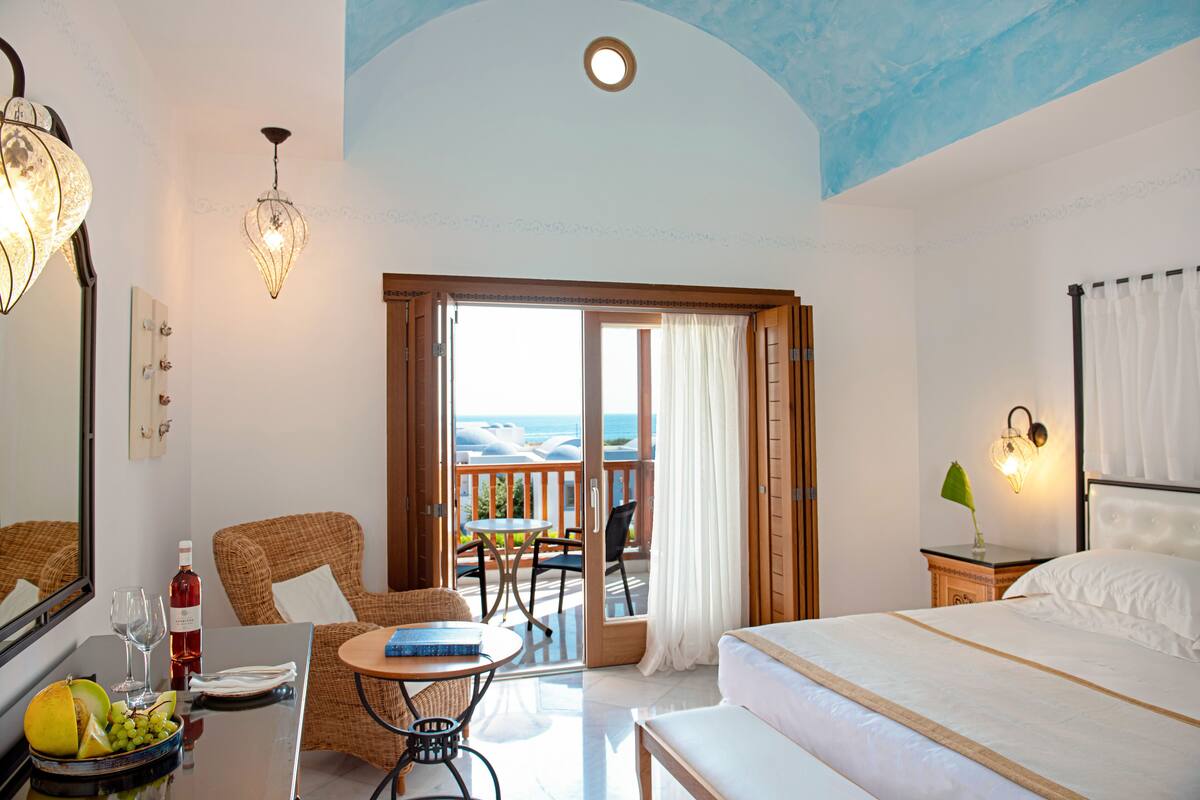 A hotel room with a bed, a dining table with chairs, a fruit bowl, a bottle of wine, and a view of the sea through the open door.