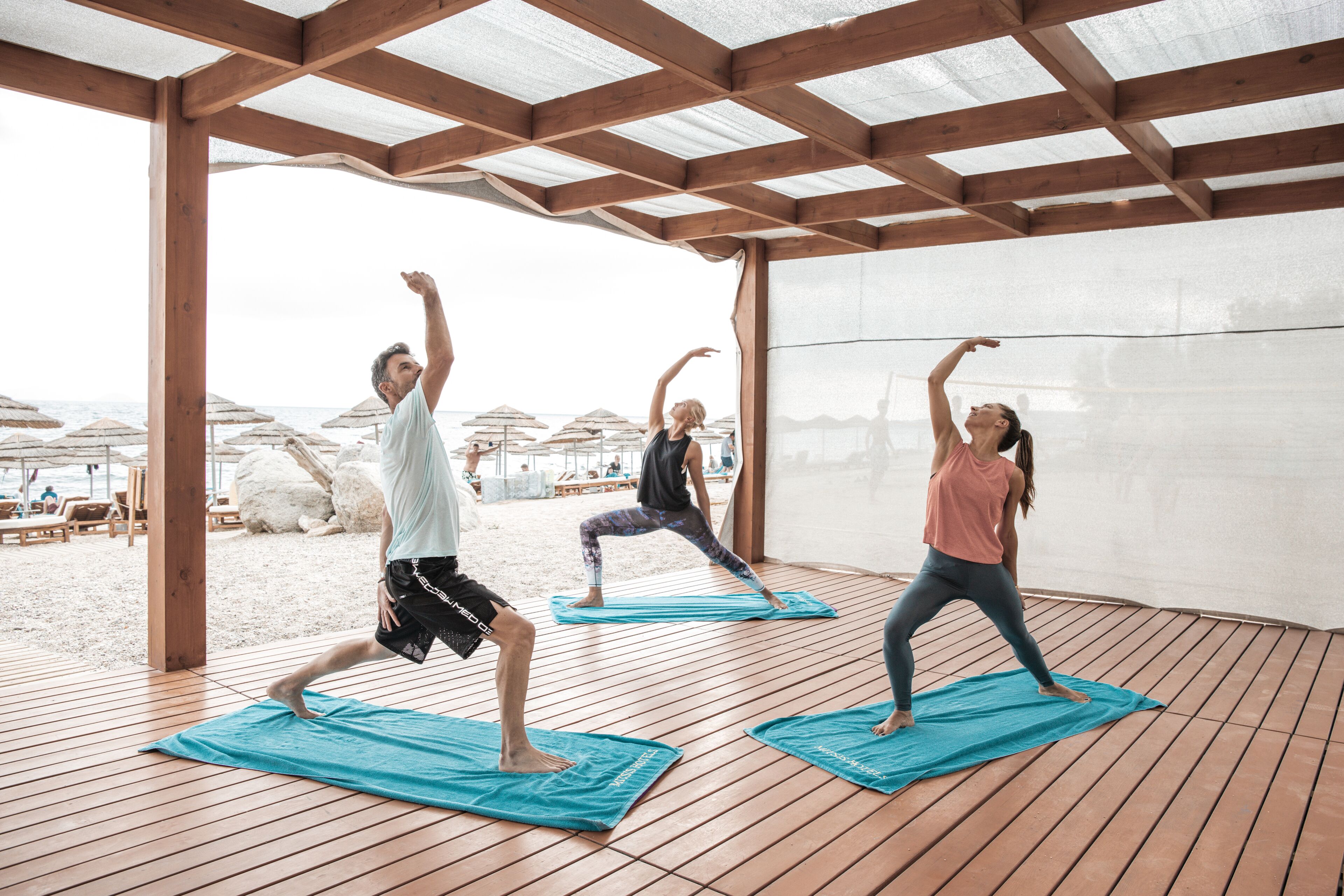 Three people practicing yoga on mats under a wooden pergola on a beach.
