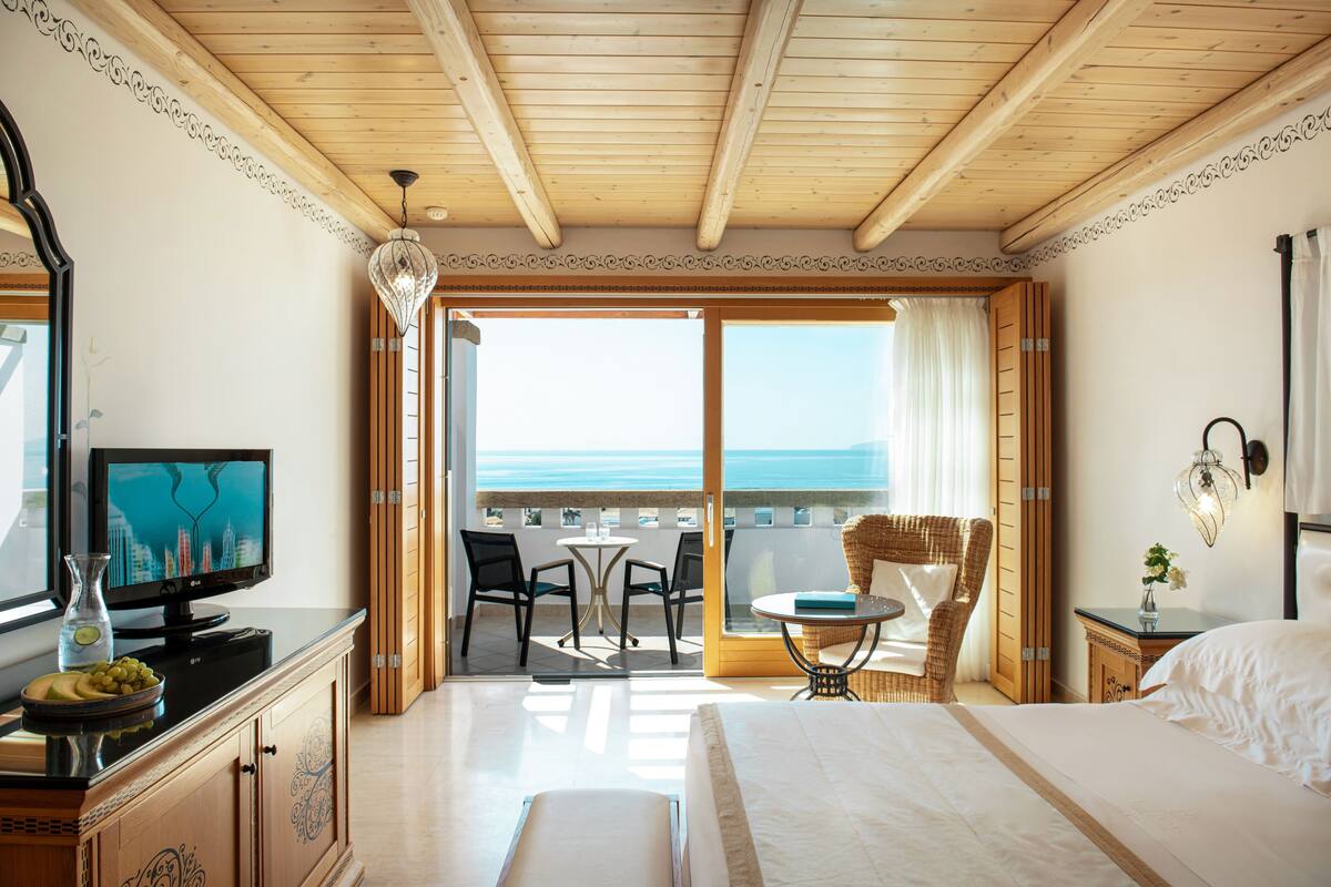 A hotel room with a bed, a television, a dining area with chairs and a table, and a view of the sea.